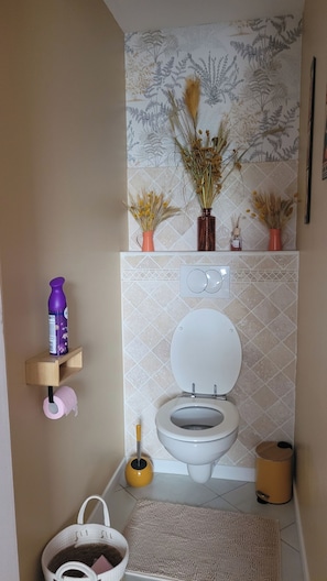 A bathroom with a toilet, a hand dryer, a trash bin, a toilet brush, and a shelf with a bottle and a roll of toilet paper. - LE CLOS DES VOYAGEURS Bed and Breakfast ALL THE COMFORTS (Saint-Christophe-de-Double)