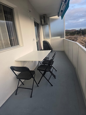 Een balkon met een witte tafel en vier zwarte stoelen, een airconditioning en uitzicht op een bouwplaats.