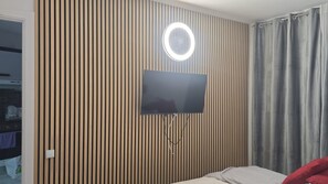 Een kamer met een televisie aan een houten lambrisering, een ronde lamp boven en een bed met een rood kussen.