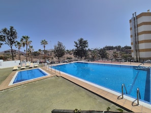 A rectangular swimming pool with a tiled deck, surrounded by palm trees and a building with a striped facade. - Sea view modern apartment in Las Americas (Costa Adeje)