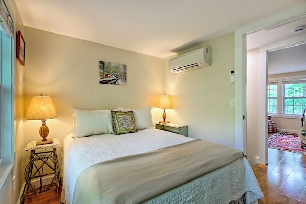 A bedroom with a bed, two bedside lamps, a wall-mounted air conditioner, and a painting. - Sunny "Charmer" Cottage w/ Folk Art & Country Primitive Style — Sleeps 4 (Lakemont)