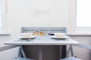 A dining table with two white bowls, a glass of water, and a small container, set against a plain wall with windows. - Earl`s Court Residence | Elegant Studio | London (Greater London)
