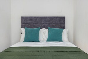 Ein Bett mit einer grünen Tagesdecke, zwei türkisfarbenen Kissen und einem grau gepolsterten Kopfteil.