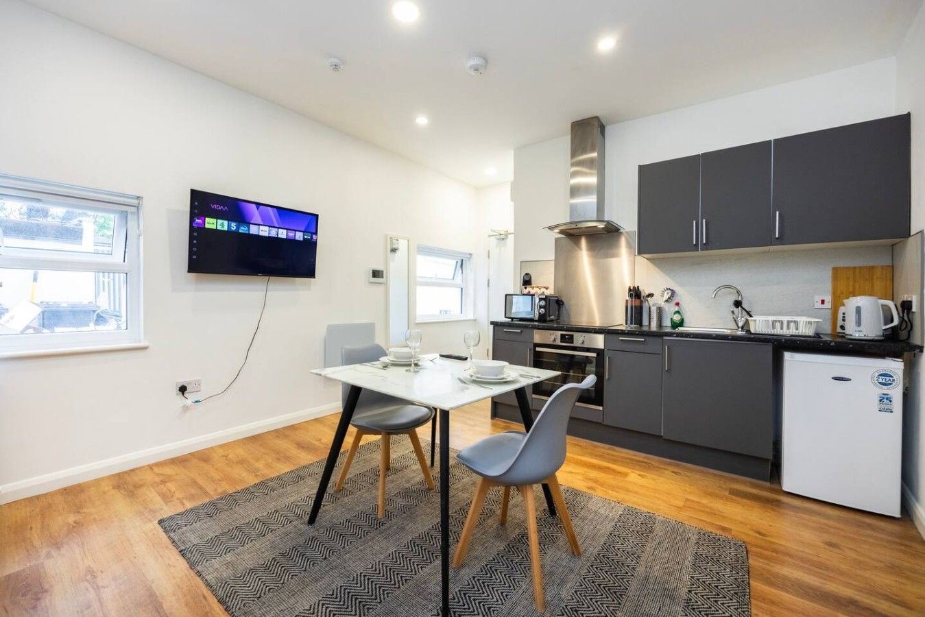 A modern kitchen with a dining area, a TV mounted on the wall, and a small refrigerator.
