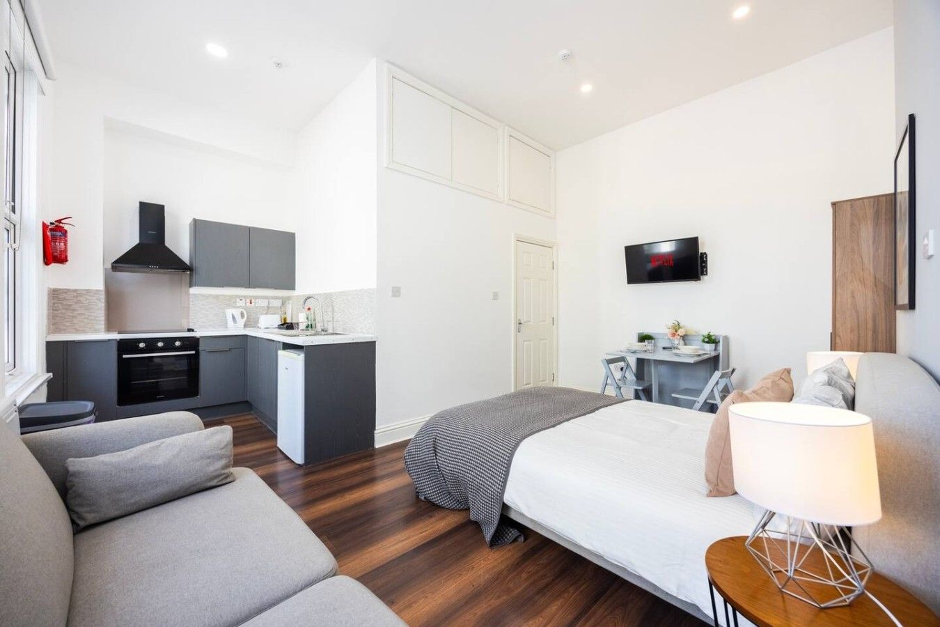 A compact hotel room with a kitchenette, a bed, a seating area, and a TV.