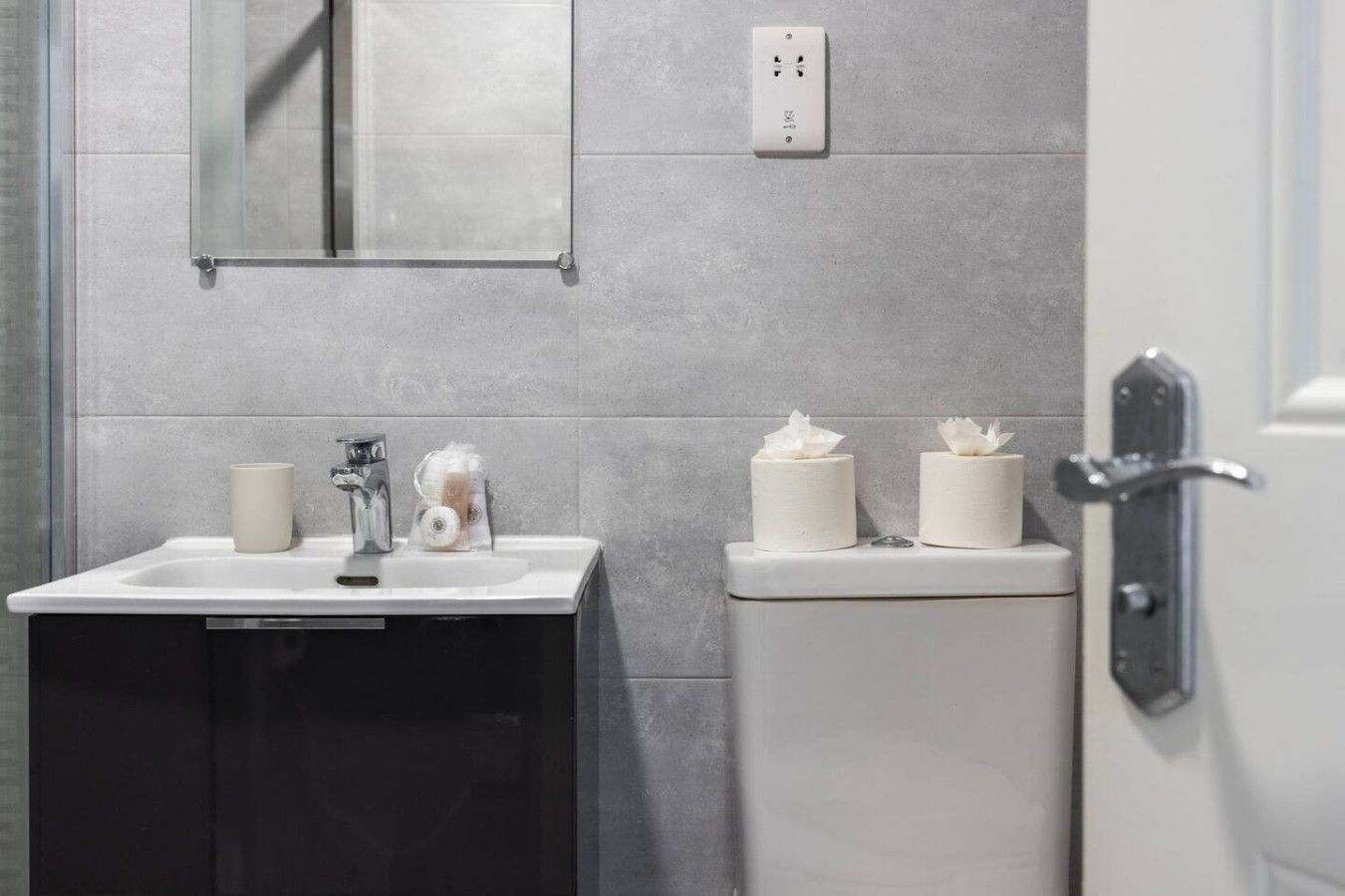 Une salle de bain avec un lavabo blanc, un miroir, des toilettes avec deux rouleaux de papier toilette et une prise électrique au mur.
