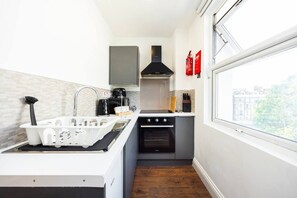 Een kleine keuken met een spoelbak, fornuis en oven, een raam met uitzicht, en een brandblusser aan de muur.