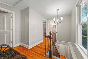 A staircase with a wooden railing leading to a second floor, a chandelier, and a view of the outdoors through a window. - Walk to Saratoga Track | 2 Kings | Pet OK (Saratoga Springs)