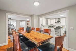 Un coin repas avec une table et des chaises en bois, une cuisine avec des meubles blancs et des appareils électroménagers, et une vue sur le coin repas à travers une porte ouverte.