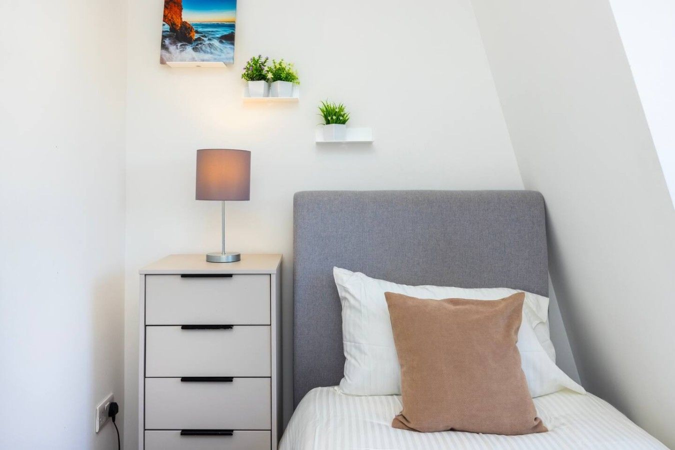 Un dormitorio con cama, una mesita de noche con lámpara, estante en la pared con plantas y un cuadro de una escena costera.