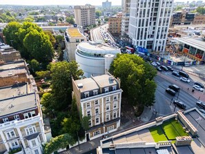 A multi-story building with a curved facade, surrounded by trees and adjacent to a busy road. - Earl`s Court Residence | Modern Studio | London (Greater London)