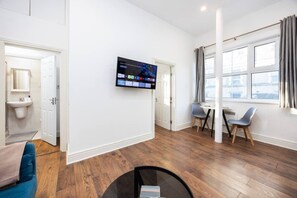 A modern hotel room with a flat-screen TV mounted on the wall, a dining area with a table and chairs, and a large window with curtains. - Earl`s Court Residence | Elegant 1BR | London (Greater London)
