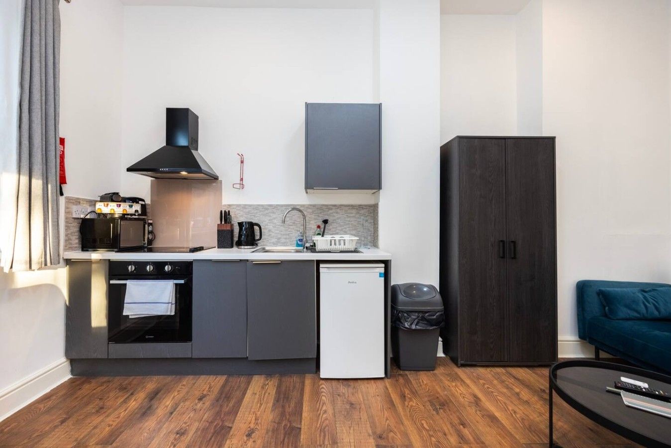 Cocina compacta con estufa, microondas y cafetera, un refrigerador pequeño y un cesto de basura.