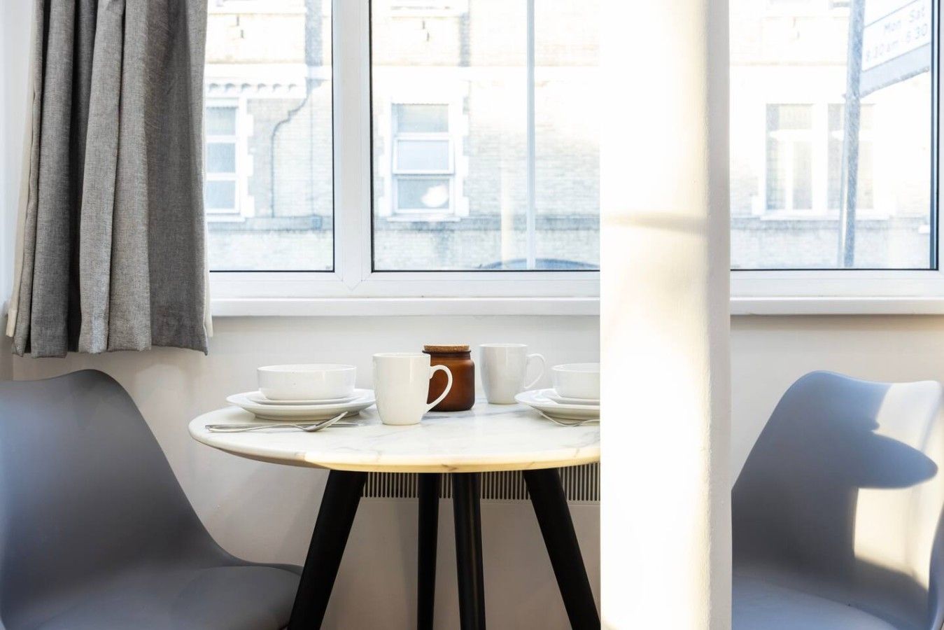 Een kamer met een gedekte tafel voor twee personen, een raam met gordijnen en uitzicht op gebouwen.