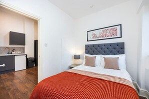 A hotel room with a bed, a nightstand, a bedside lamp, a framed picture on the wall, and a view of the adjacent kitchen area. - Earl`s Court Residence | Elegant 1BR | London (Greater London)
