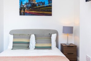 A neatly made bed with patterned pillows, a bedside lamp, and a framed picture of a cityscape with lights on a wall. - Earl`s Court Residence | Elegant Studio | London (Greater London)