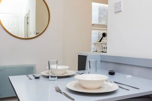 A clean, modern dining area with a white table set for two, including plates, bowls, glasses, and cutlery. - Earl`s Court Residence | Elegant Studio | London (Greater London)