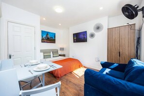 A compact hotel room with a bed, a dining table with chairs, a blue sofa, a wardrobe, and a wall-mounted fan. - Earl`s Court Residence | Elegant Studio | London (Greater London)