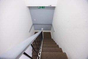 A staircase with a fire exit sign above, a metal handrail, and carpeted steps. - Earl`s Court Residence | Elegant Studio | London (Greater London)