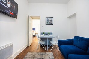 A compact living space with a dining area, a blue sofa, and a television mounted on the wall. - Earl`s Court Residence | Stylish 1BR | London (Greater London)