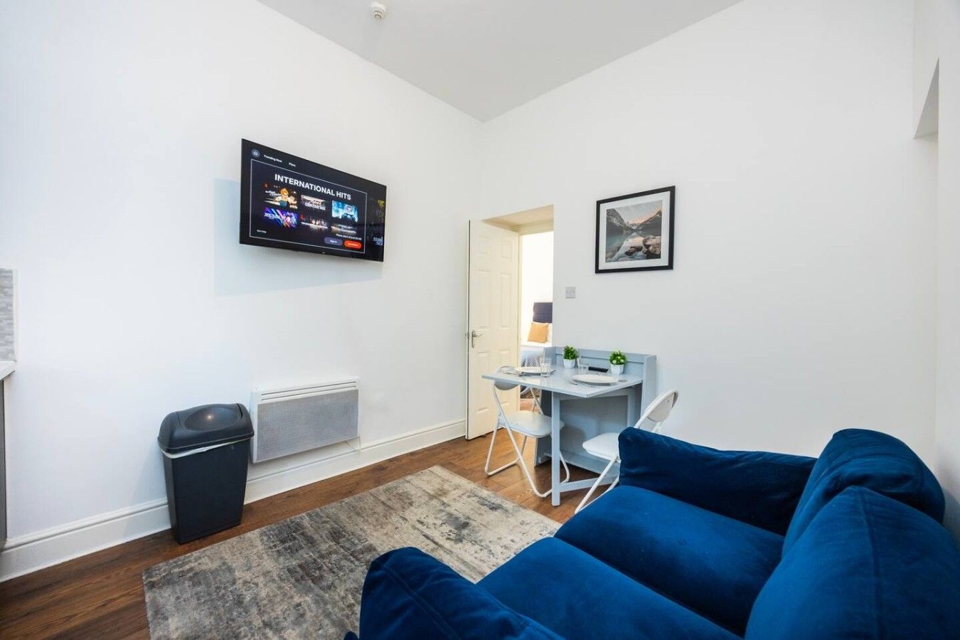 Un salon moderne avec un canapé bleu, un téléviseur à écran plat diffusant des « succès internationaux », une petite table à manger avec des chaises et un tableau encadré accroché au mur.