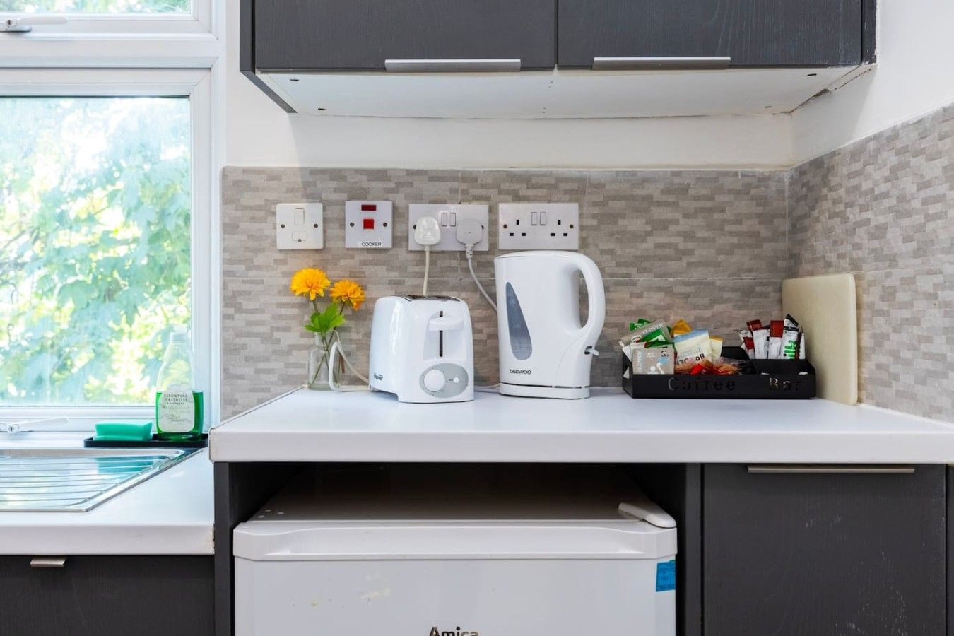 Een keuken met een broodrooster, waterkoker en koffiezetapparaat op het aanrecht, een koffiemachine, en een doos koffiecups.