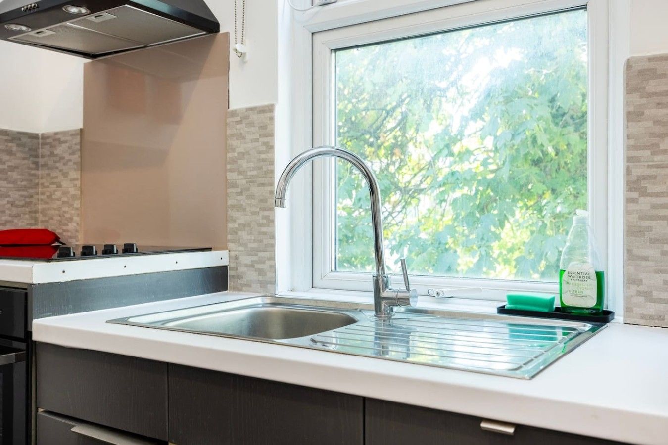 A kitchen with a stainless steel sink, a window with a view of greenery, and a red appliance on the counter.