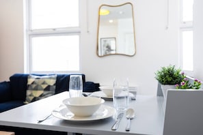 A dining table with a bowl, glass, and cutlery, a sofa with a geometric-patterned cushion, a mirror on the wall, and a potted plant with flowers. - Earl`s Court Residence | Stylish Studio | London (Greater London)