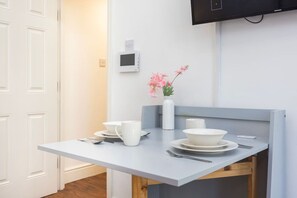 A compact dining area with a table, white plates, cups, and a vase with pink flowers. - Earl`s Court Residence | Elegant Studio | London (Greater London)