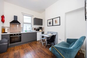 A compact kitchen with a stove, oven, and modern appliances. There is a dining area with a table and chairs, and a blue chair in the foreground. - Earl`s Court Residence | Elegant 1BR | London (Greater London)