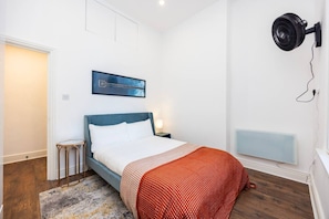Ein Schlafzimmer mit einem Bett, einem Nachttisch, einem Wandventilator, einer Heizung und einem gerahmten Bild an der Wand.