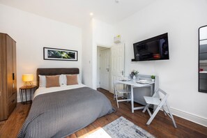 Ein Schlafzimmer mit Bett, Nachttisch, Lampe, Fernseher, Schreibtisch, Stuhl und einer Tür zu einem anderen Raum.