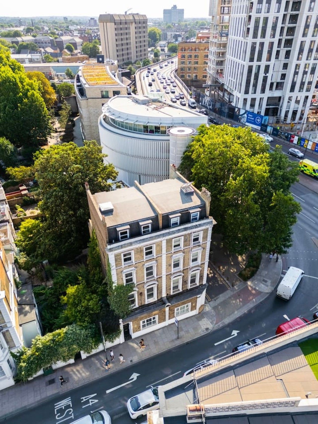 Un edificio de varios pisos con fachada curva, rodeado de árboles y al lado de una calle con mucho tráfico.