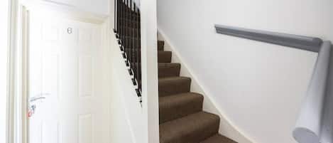 Un escalier avec des marches recouvertes d’une moquette marron, une porte blanche portant un numéro et une main courante en métal.