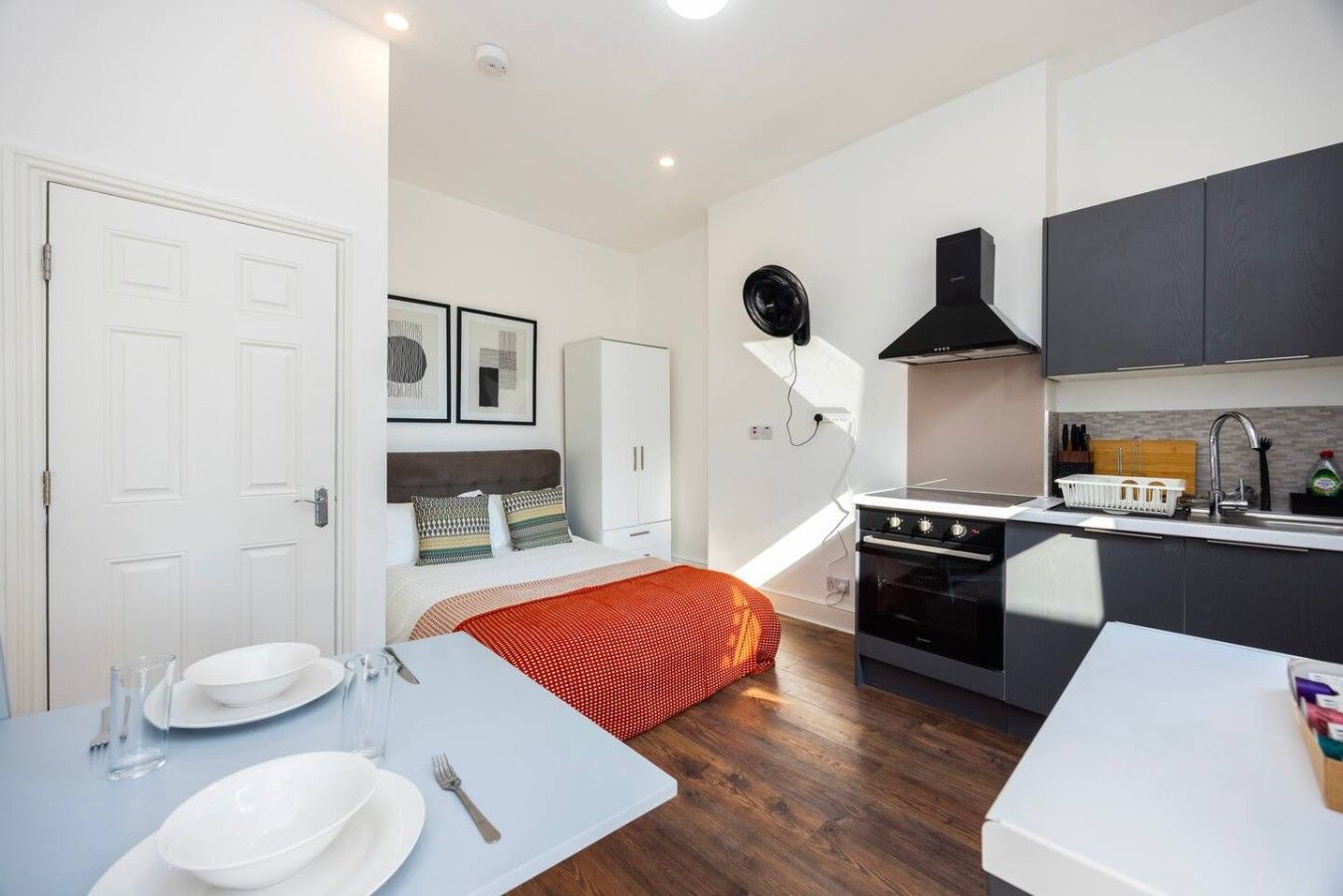 Habitación de hotel compacta con kitchenette, una cama con manta roja, una zona de comedor con mesa preparada para dos personas y un armario blanco.