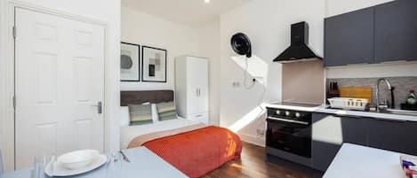 Habitación de hotel compacta con kitchenette, una cama con manta roja, una zona de comedor con mesa preparada para dos personas y un armario blanco.