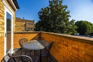 Ein Balkon mit rundem Tisch und Stühlen, einer Backsteinmauer und Blick auf einen Baum und ein Gebäude.