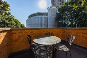 Een terras buiten met een ronde tafel en rieten stoelen, een bakstenen muur en een modern gebouw op de achtergrond.