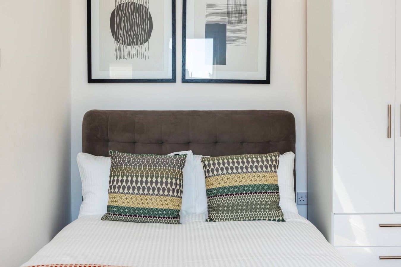 A neatly made bed with patterned pillows, a tufted headboard, and a white wardrobe with gold handles.