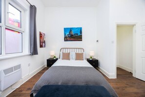 A bedroom with a bed, two bedside tables, a window with curtains, a radiator, and a wall-mounted painting. - Earl`s Court Residence | Stylish Studio | London (Greater London)