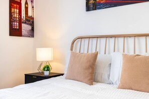 Ein ordentlich bezogenes Bett mit weißer Bettwäsche und zwei Kissen, ein Nachttisch mit Lampe und einer kleinen Pflanze sowie ein an der Wand hängendes Stadtbild.
