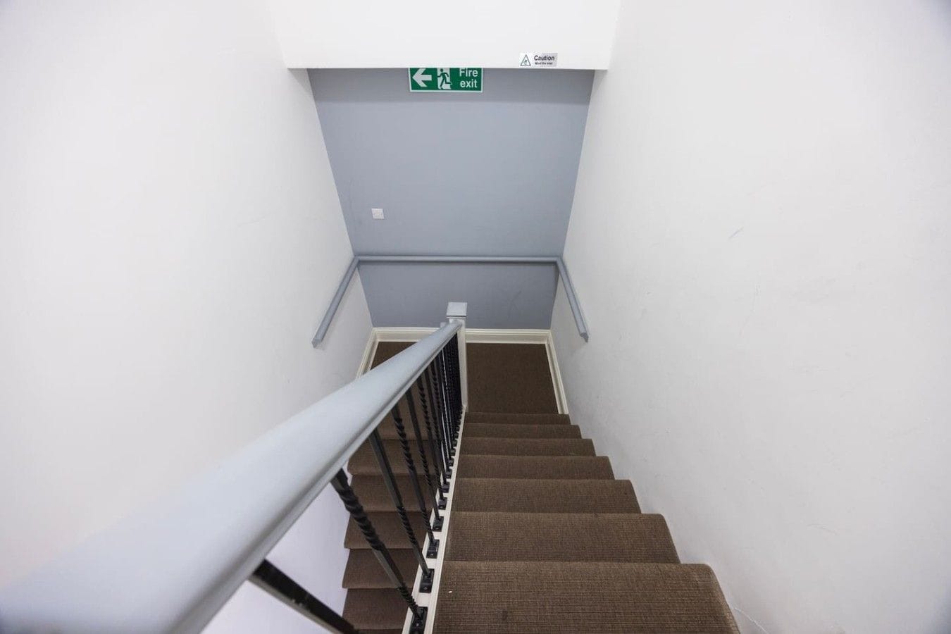 A staircase with a fire exit sign above, a metal handrail, and carpeted steps.