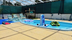 En swimmingpool med oppustelige legetøj, liggestole og et overdækket område med en grøn presenning.