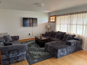 Uma sala de estar com sofá seccional, televisão de tela plana na parede, mesa lateral pequena e janela com cortinas leves.