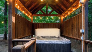 Een hottub in een houten tuinhuisje, omgeven door lichtjes, een houten hek en bomen.