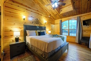 A wooden cabin bedroom with a bed, nightstands, a ceiling fan, and a large window. - Private & Rustic | 2 Kings + Hot Tub + Firepit (Broken Bow)
