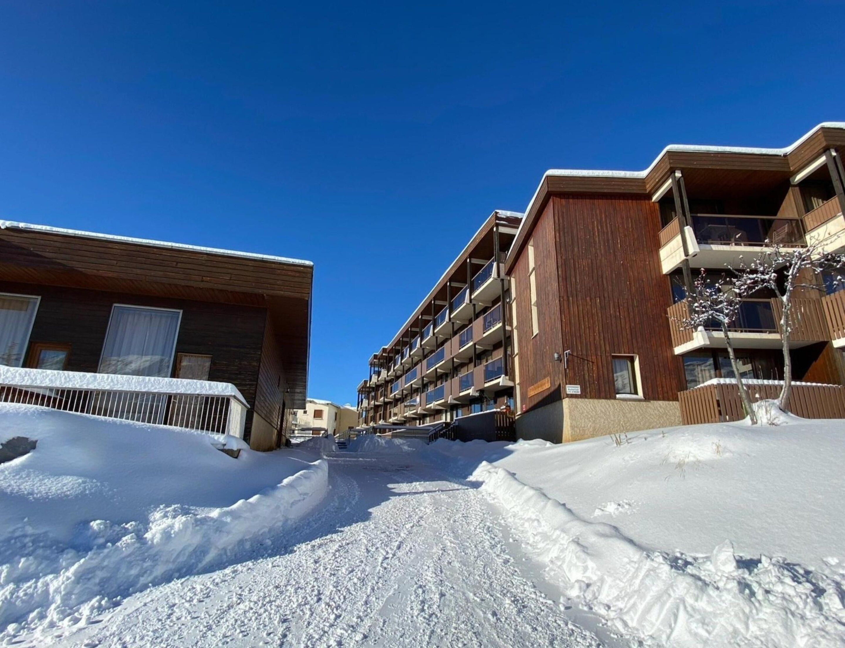 A snow-covered street with modern buildings, including a multi-story structure with balconies.