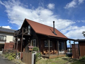 A wooden cabin with a sloped roof and a chimney, featuring a porch with chairs and a small garden area.