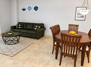 Ein Wohnzimmer mit einem grünen Sofa, einem Holzespeisetisch mit Stühlen, einem kleinen Beistelltisch mit einer Pflanze und einem gemusterten Teppich.