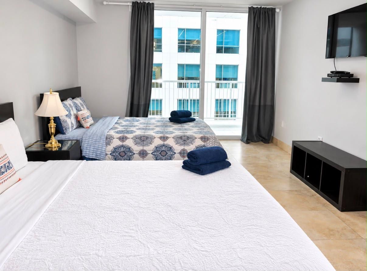 A hotel room with a bed, a nightstand, a lamp, a TV, and a view of the outside.